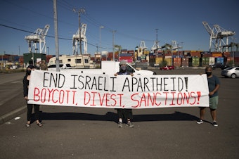 caption: Activists and allies of the Palestinian feminist organization Falastiniyat hold a sign calling for the end to Israeli apartheid while others block an intersection in protest of the Israeli Zim San Diego Vessel, on Thursday, June 17, 2021, at the Port of Seattle. 