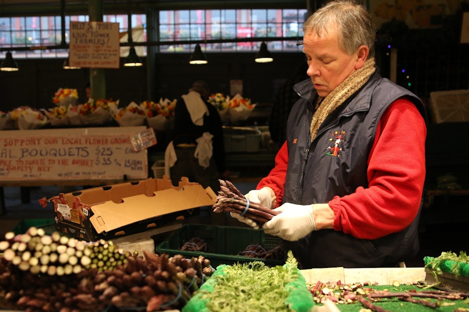 caption: Mike Osborn at Sosio's Fruit and Produce in the Pike Place Market on March 25, 2020. Behind him and across the aisle are flower bouquets he's selling for flower vendors that can't legally open.