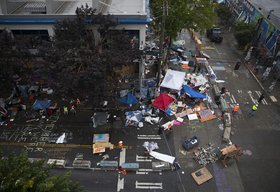 caption: Seattle Department of Transportation employees remove items from outside of the Seattle Police Department's East Precinct building after the Capitol Hill Organized Protest zone was cleared by Seattle Police Department officers early Wednesday morning, July 1, 2020, at the intersection of 12th Avenue and East Pine Street in Seattle. 