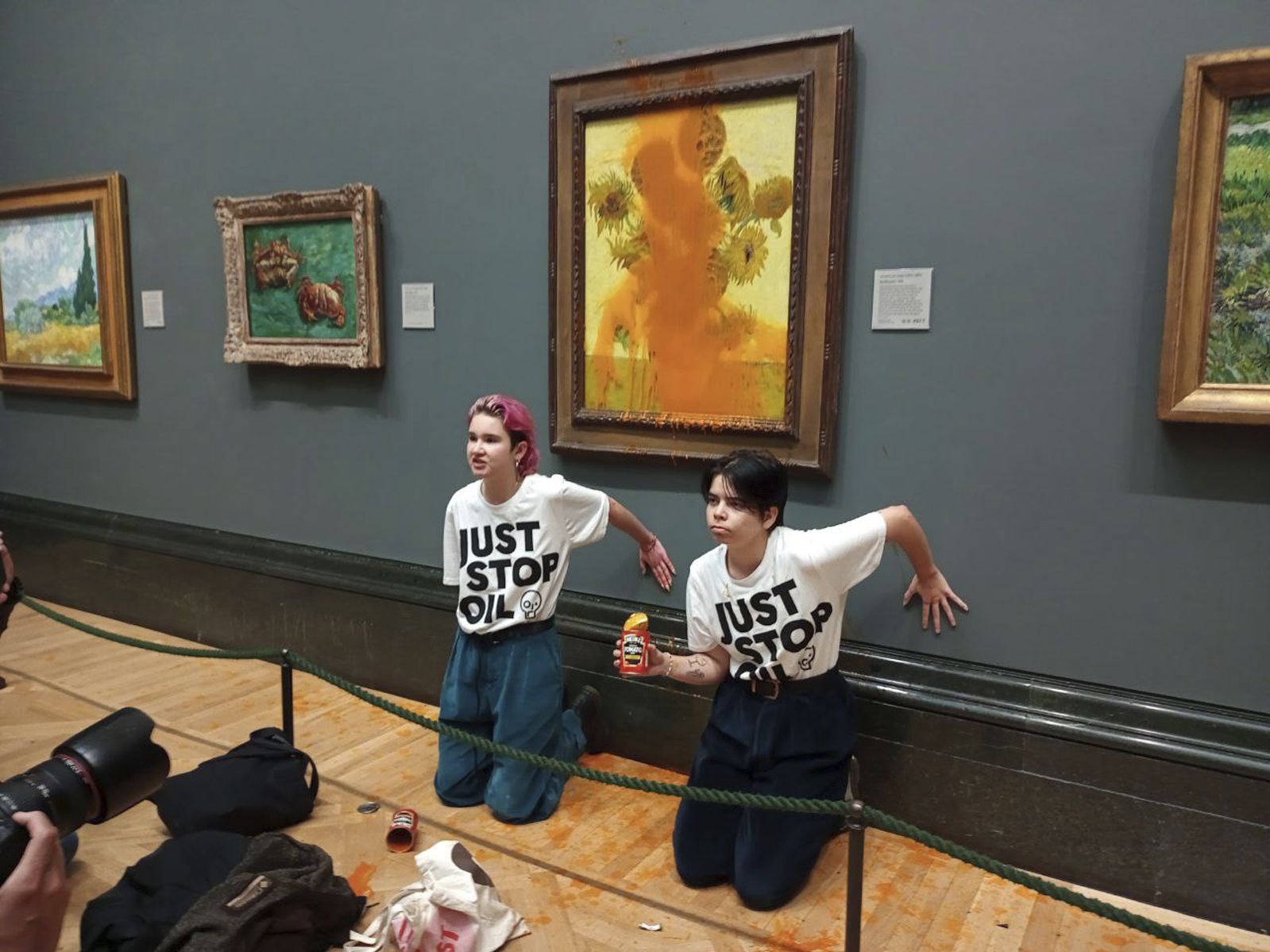 caption: A handout photo issued by the group Just Stop Oil shows two protesters who threw soup at Vincent Van Gogh's famous 1888 work <em>Sunflowers</em> at the National Gallery in London on Friday.