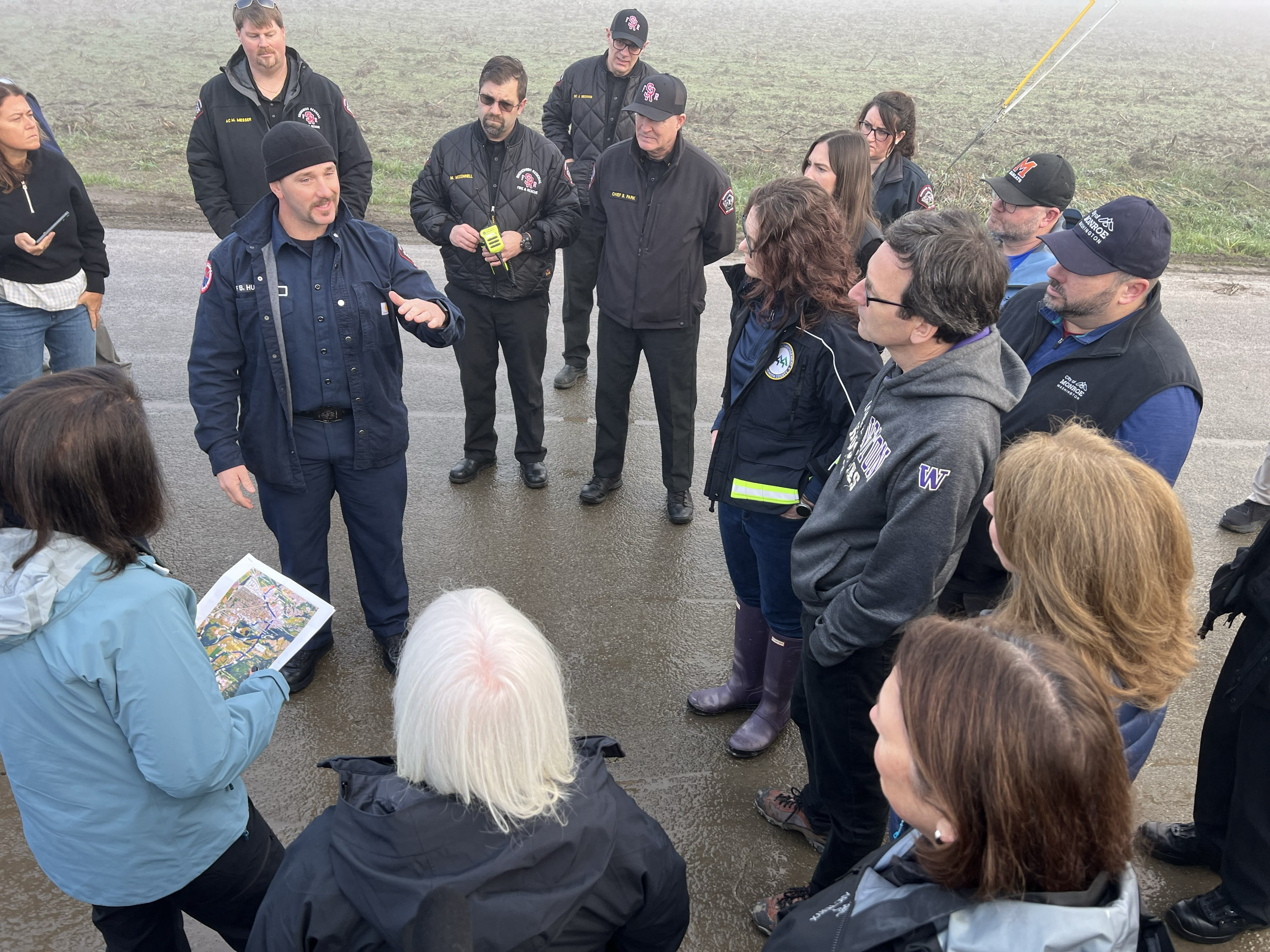 caption: Washington state Gov. Bob Ferguson, U.S. Sen. Patty Murray (D-WA), U.S. Rep. Suzan DelBene (D-WA), and U.S. Rep. Kim Schrier (D-WA) stand by a soggy field in Monroe, Wash., as firefighter Brandon Huber describes recent flood rescues undertaken in this area.