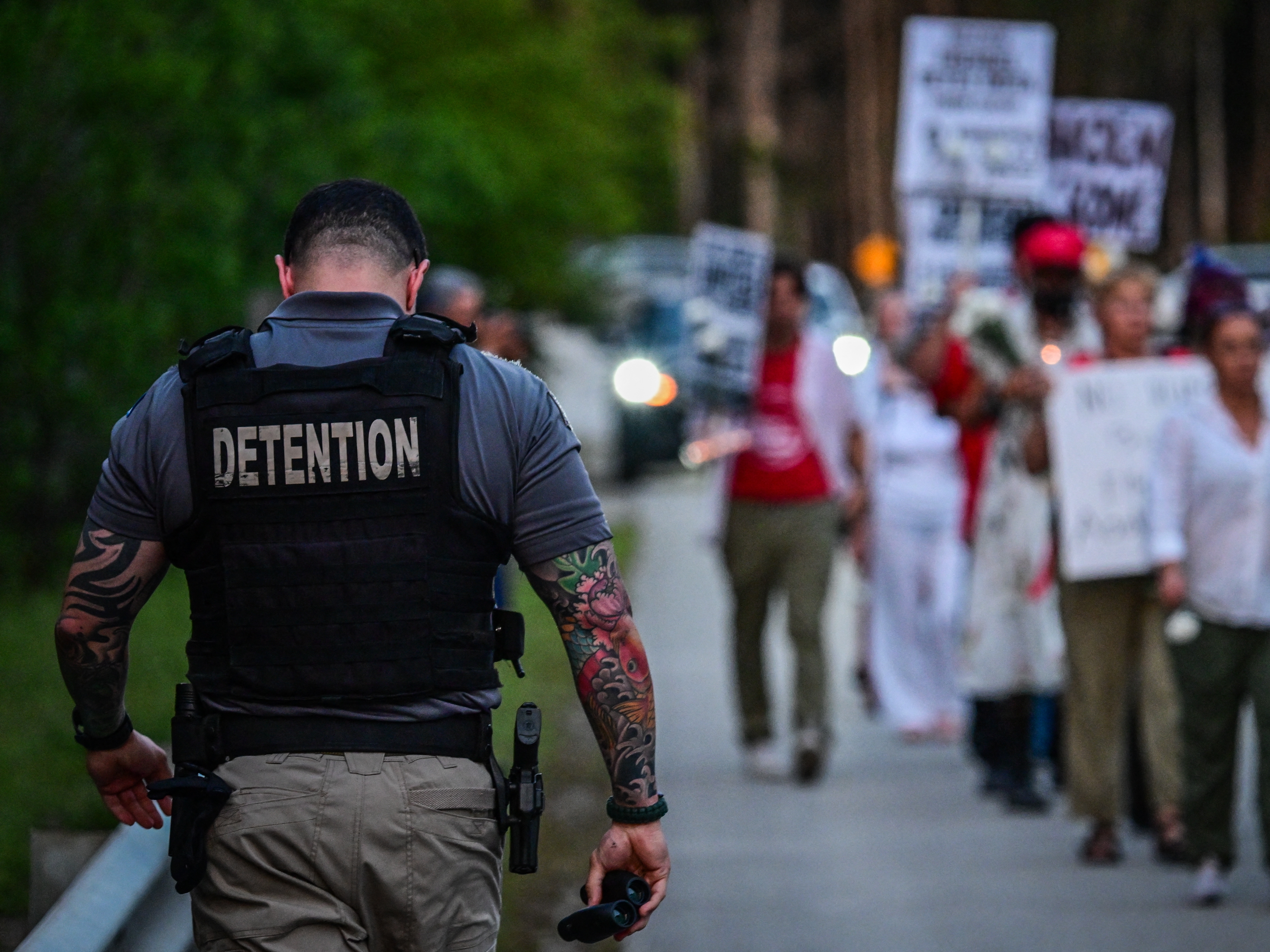 caption: A corrections officer walks beside people holding candles, signs, and flowers during a vigil outside the Krome Detention Center in Miami in May 2025, protesting U.S. Immigration and Customs Enforcement custody and mass deportations.