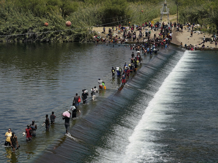 caption: In this Sept. 18, 2021, file photo Haitian migrants use a dam to cross into the United States from Mexico in Del Rio, Texas.