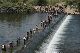 caption: In this Sept. 18, 2021, file photo Haitian migrants use a dam to cross into the United States from Mexico in Del Rio, Texas.