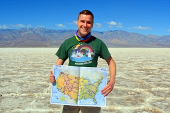 caption: Mikah Meyer holds up a map of his journey at his 313th stop: Badwater Basin in Death Valley National Park, the lowest point in North America. (Courtesy of Mikah Meyer)