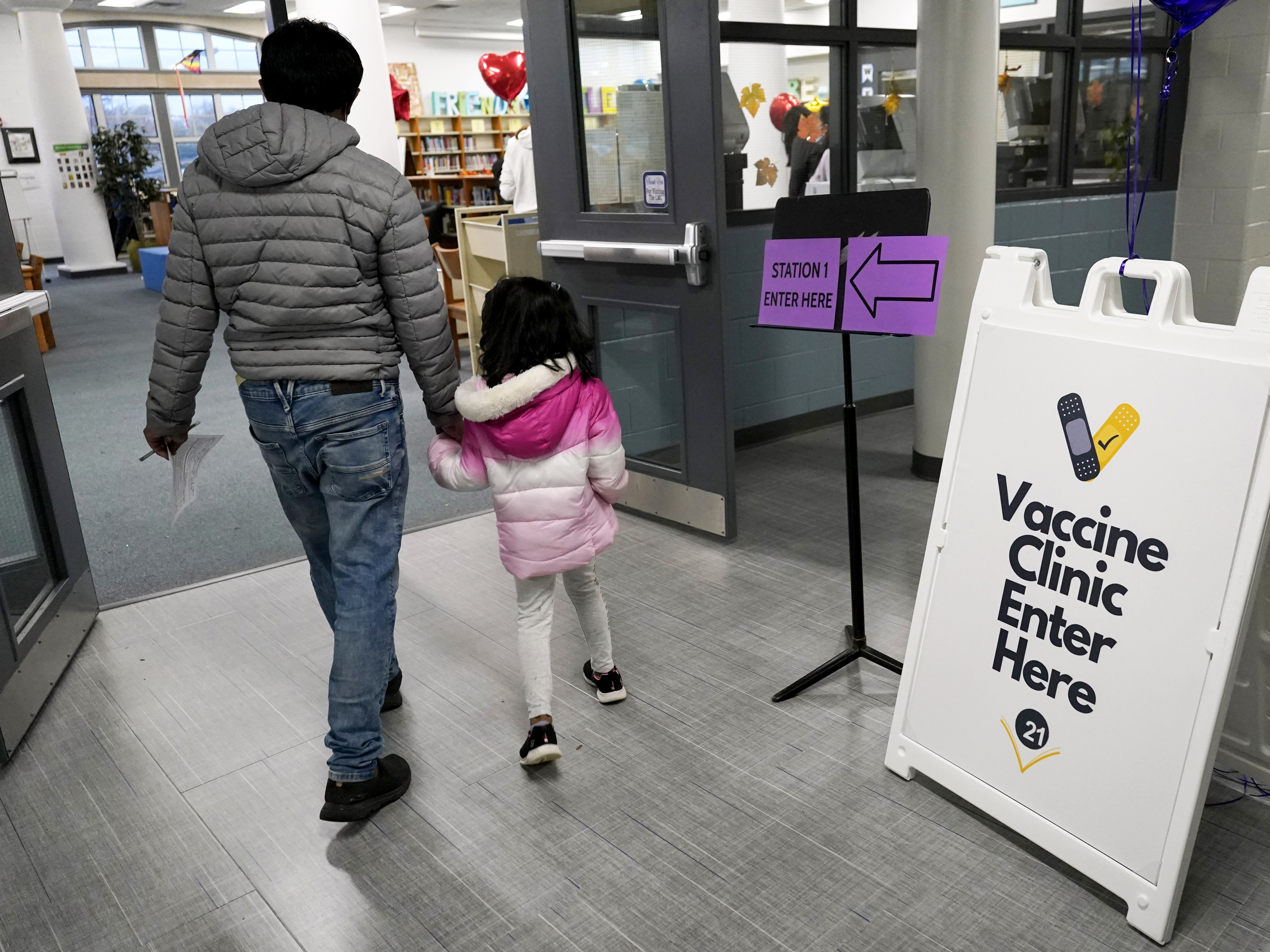 caption: An information sign is displayed as a child arrives with her parent to receive the Pfizer COVID-19 vaccine for children 5 to 11-years-old at London Middle School in Wheeling, Ill., last month.