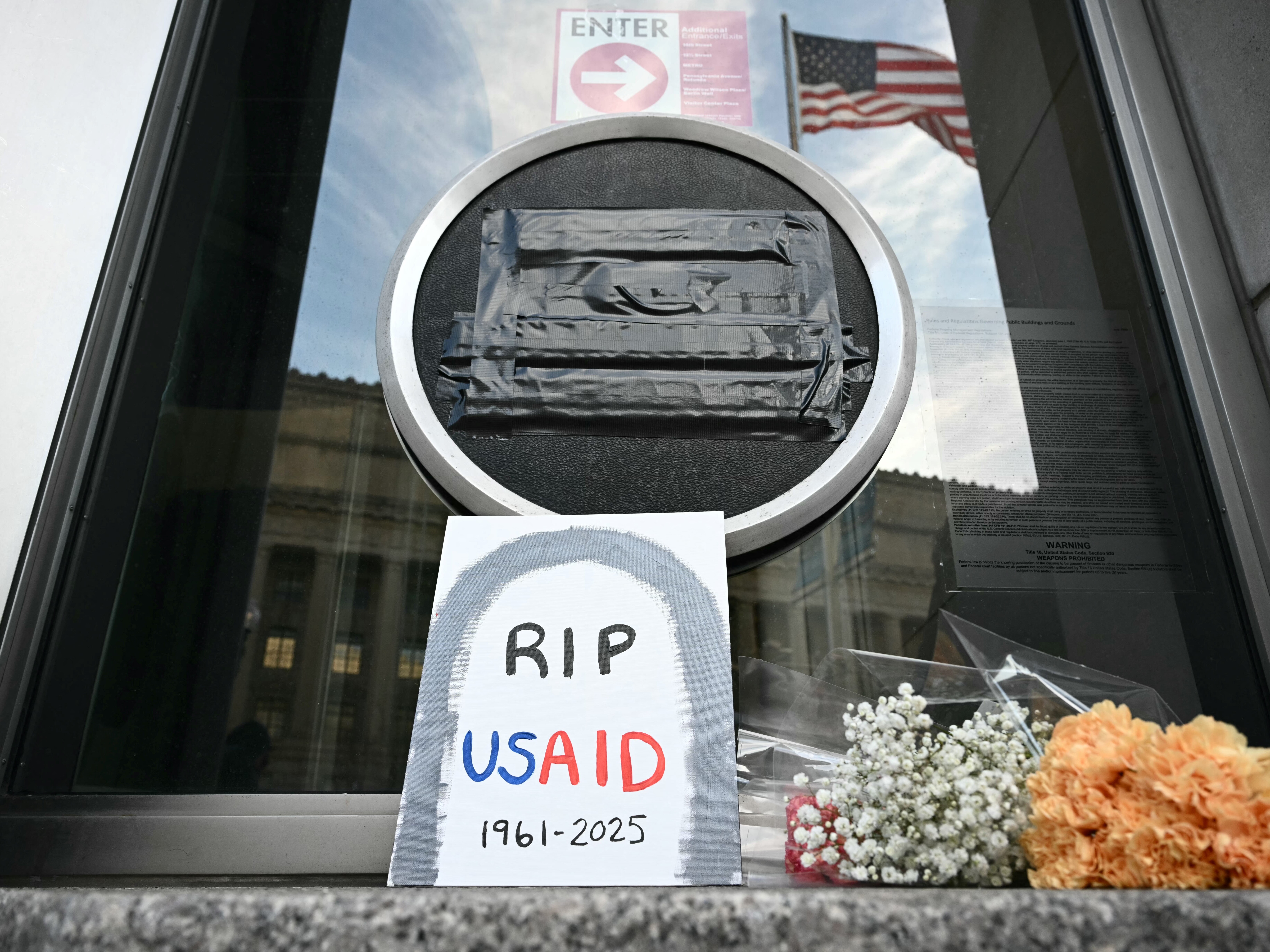 caption: Tributes are placed beneath the covered seal of the United States Agency for International Development (USAID) at their headquarters in Washington, D.C., on February 7, the day that President Donald Trump called for the agency to be shuttered. July 1 marks the agency's official demise.