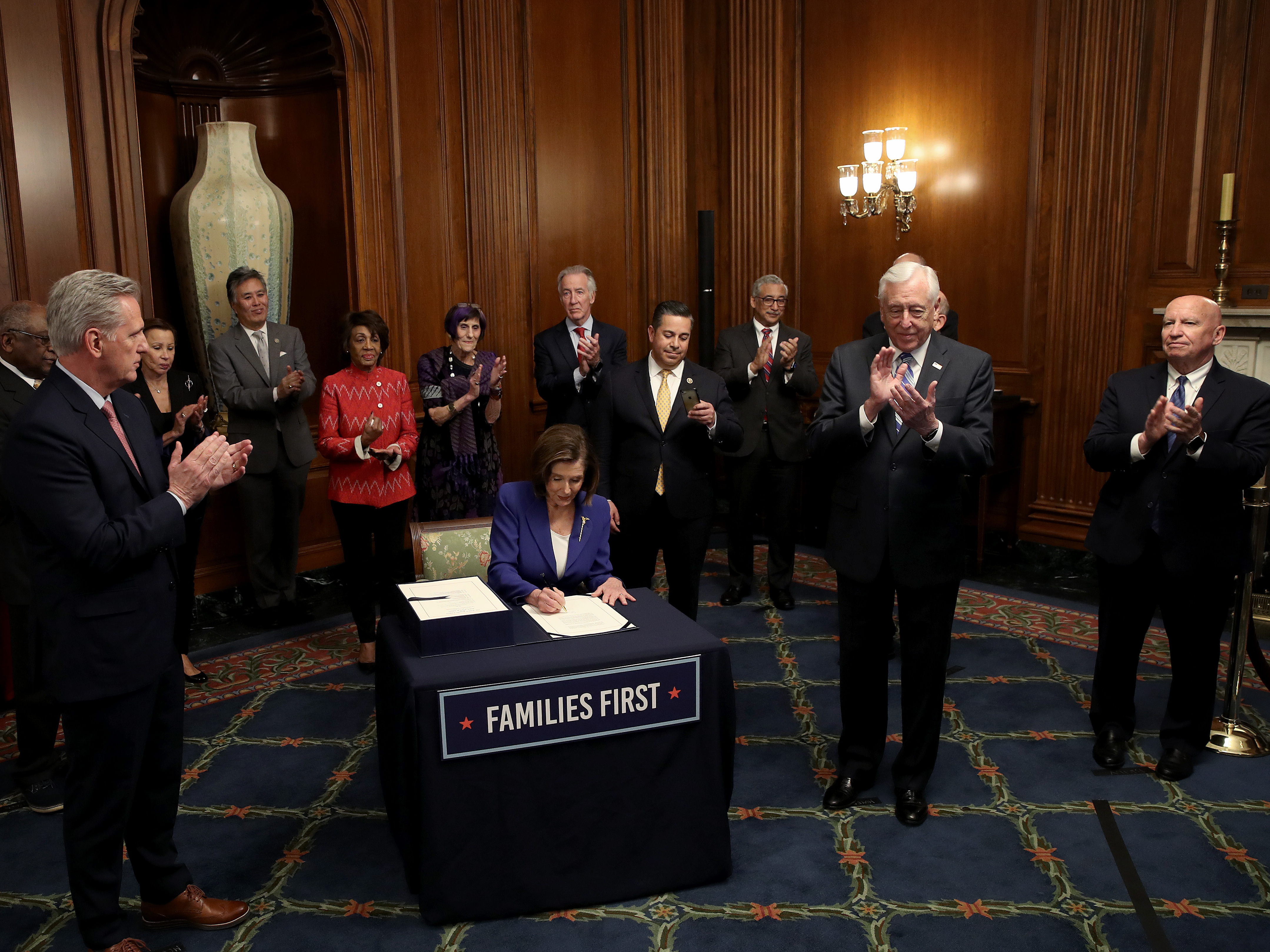 caption: House Speaker Nancy Pelosi, surrounded by a bipartisan group of members of the House, signs the CARES Act on March 27.