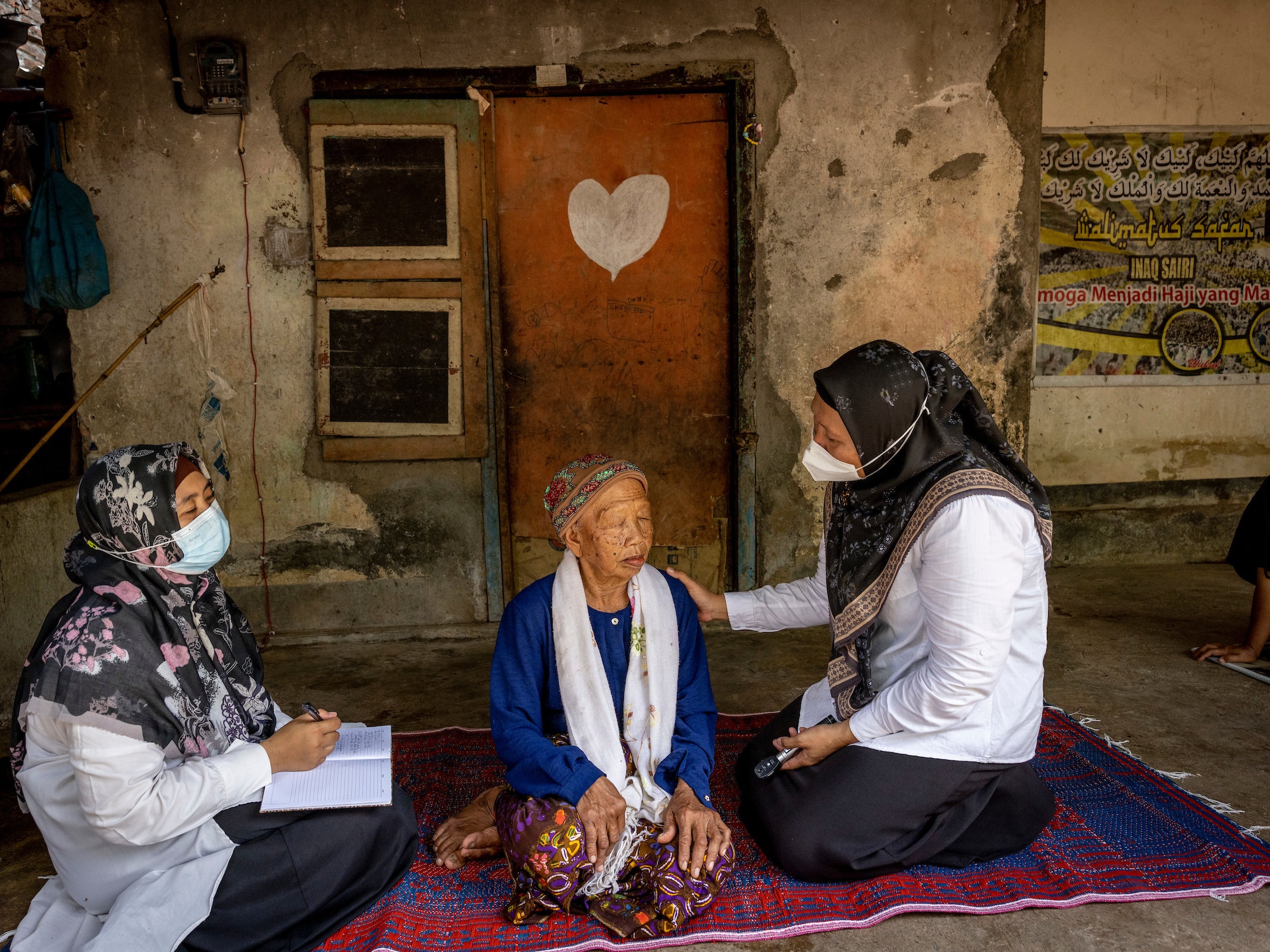 caption: Two health workers see a patient.  “Cataract cut my main income source," the patient said before she had surgery. "I cannot sell anything, I cannot write anything. It’s difficult to continue with my job.”  