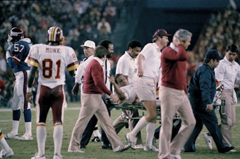 caption: Washington Redskins quarterback Joe Theismann gestures as he is carried off the field at RFK Stadium in Washington, D.C., Nov. 18, 1985. Theismann injured his right leg during second quarter action. 