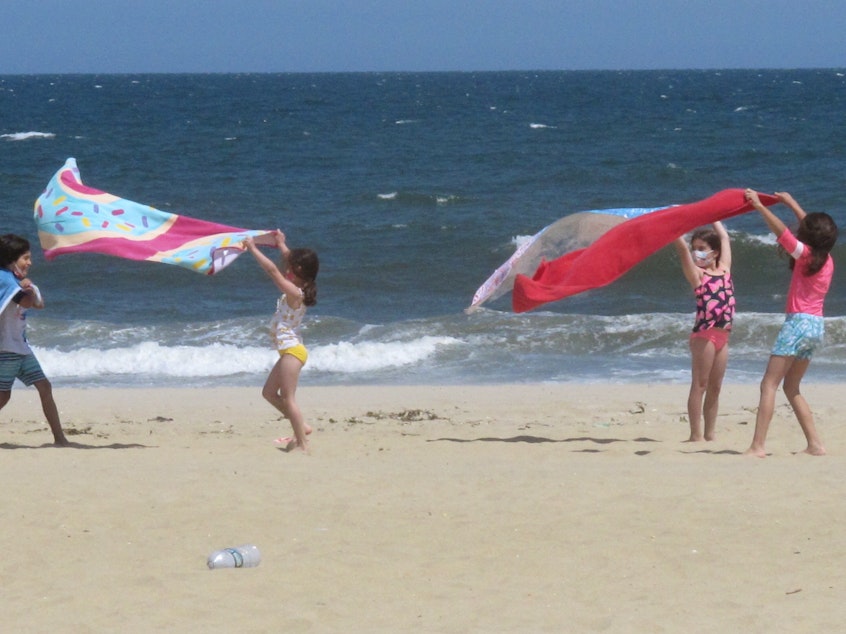 caption: Children play with beach towels on a windy day in Belmar, N.J., on Tuesday. Millions are expected to hit the road or board a plane to celebrate Memorial Day weekend as more people get vaccinated and COVID-19 restrictions are scaled back.