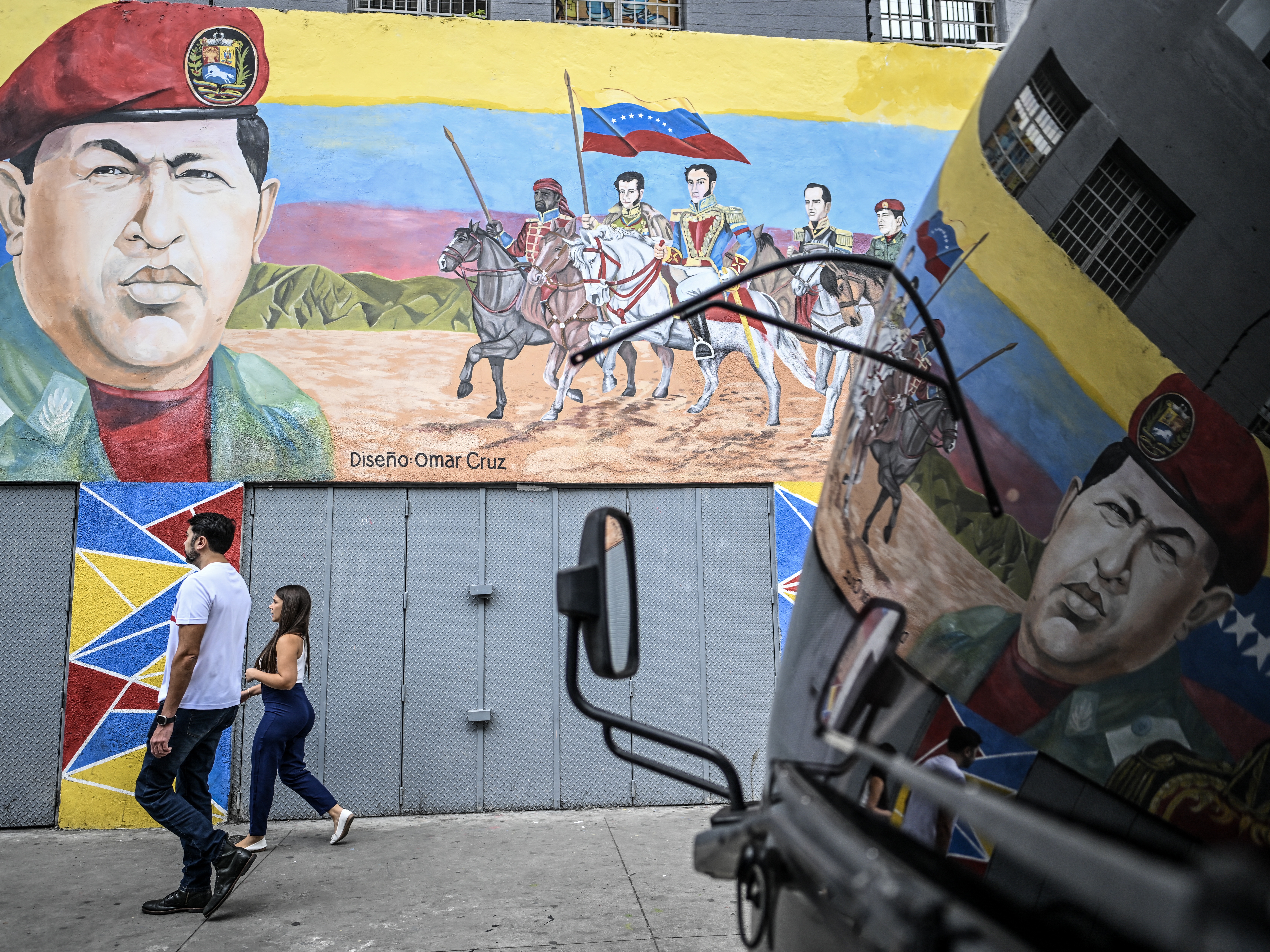 caption: People walk past a mural depicting former Venezuelan president Hugo Chavez alongside Venezuelan independence heroes in Caracas on Dec.17, 2025.