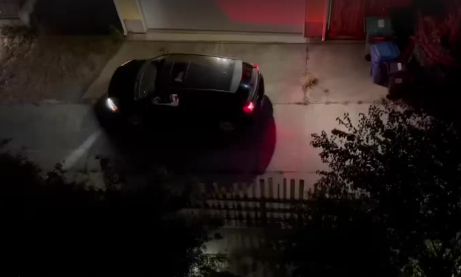 caption: A still from a video of a car pulling up to the back of Janus Apartments in Greenwood, in north Seattle, late summer 2022. In this video, the driver peels off what appears to be cash from a wad and hands it to a woman.