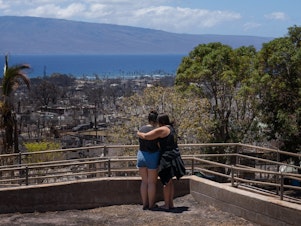 caption: Two women embrace and cry as they look out over Lahaina, in Maui, Hawaii, which was severely damaged by a wildfire in August.