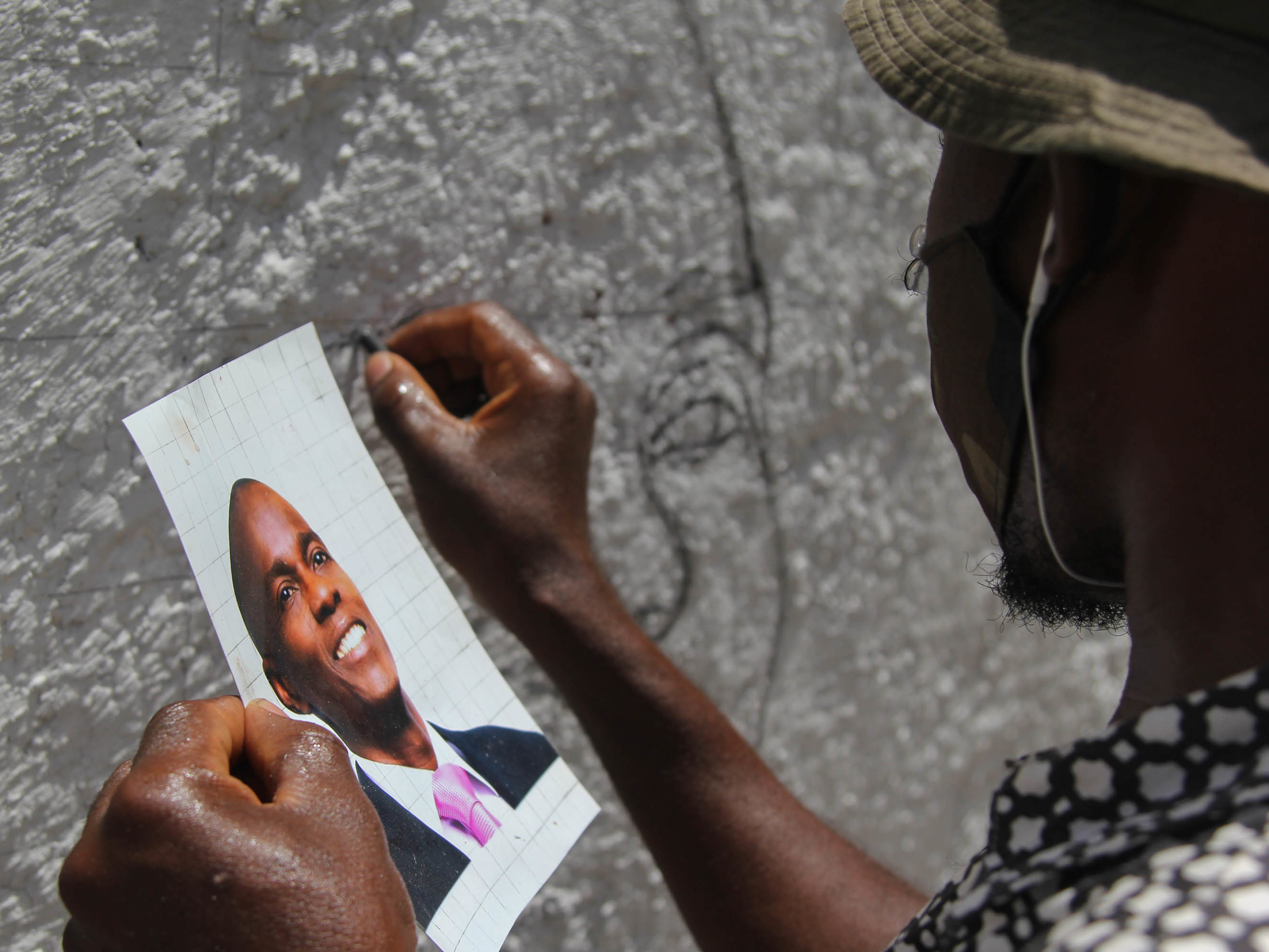 caption: An artist who goes by the name Jamesy Jay paints a mural of slain Haitian president Jovenel Moïse on the road leading to the president's private residence above Port-au-Prince.