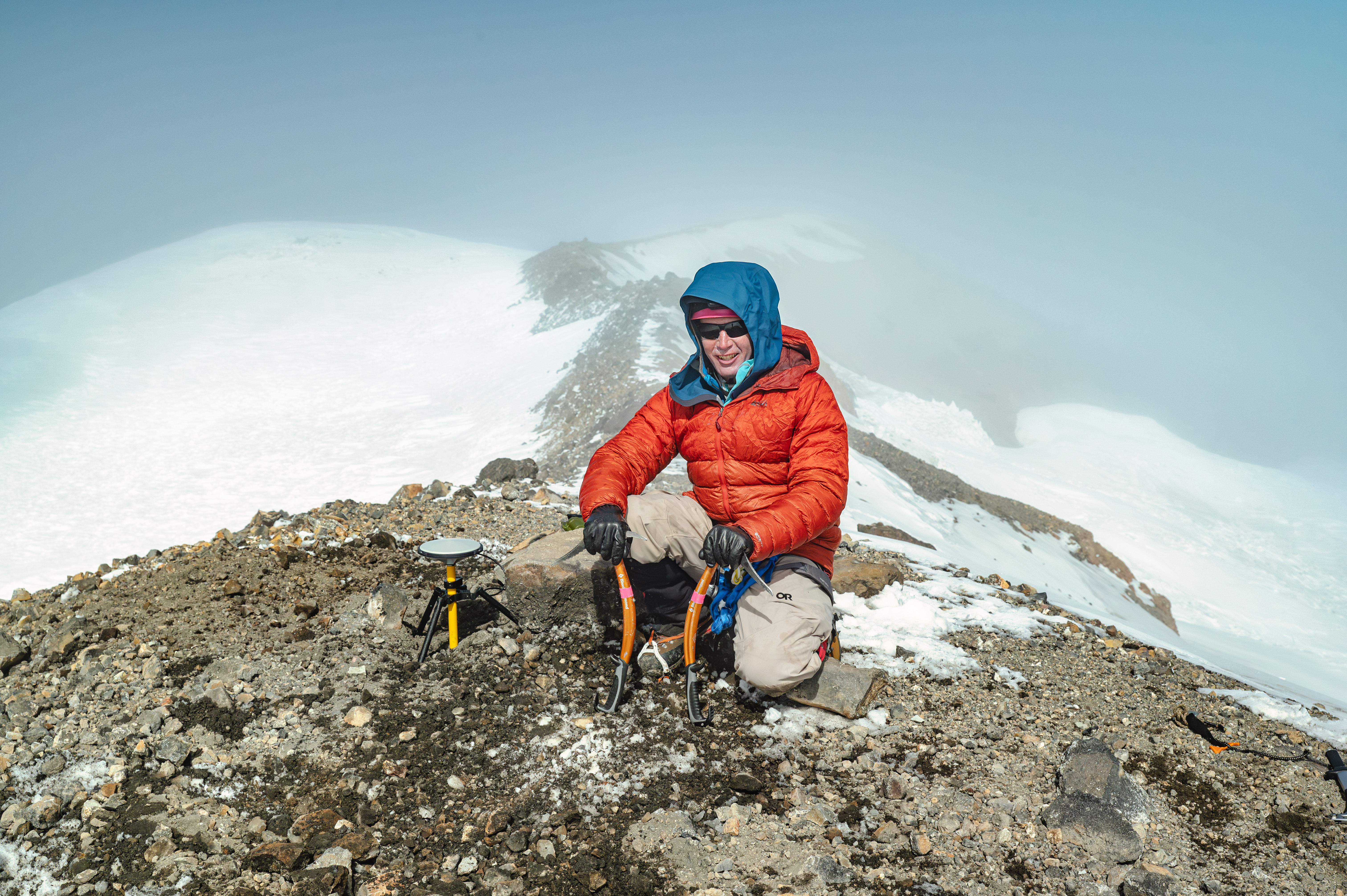 caption: Eric Gilbertson of Seattle University crouches next to a surveying device on the southwest rim of Mount Rainier on Sept. 21, 2024, with Columbia Crest in the background.