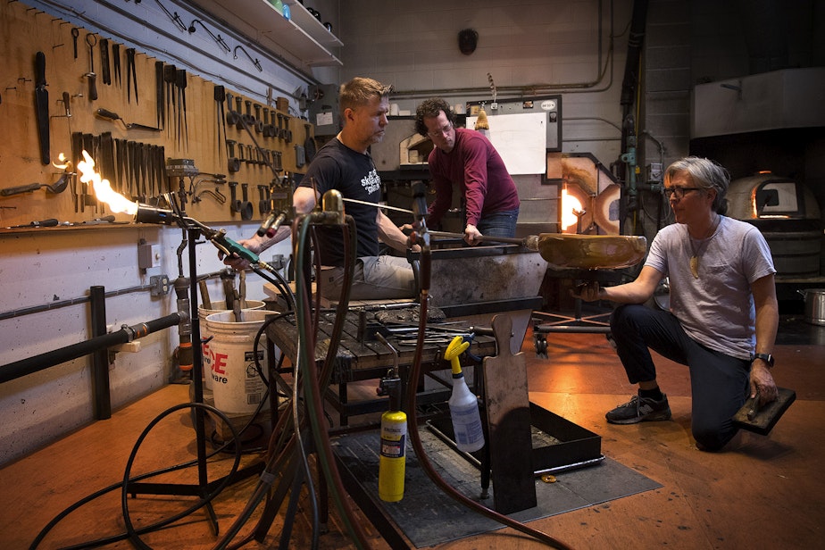 caption: Assistants Janusz Pozniak, left, and Sean Albert, center, assist glass artist Preston Singletary, right, on Tuesday, December 11, 2018, as he sculpts a form to later do design work on at his studio in Seattle.