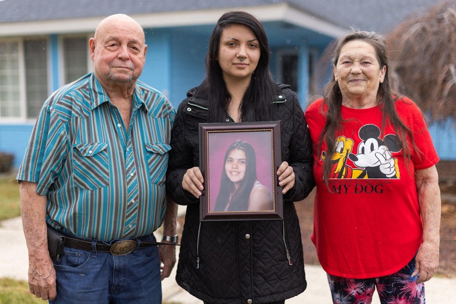 caption: From left, Charlie, Charlotte, and Bonnie Groo stand outside the home where Kit Nelson-Mora was raised in Yakima, Washington. Charlotte holds a photo of Kit, who went missing at age 16.