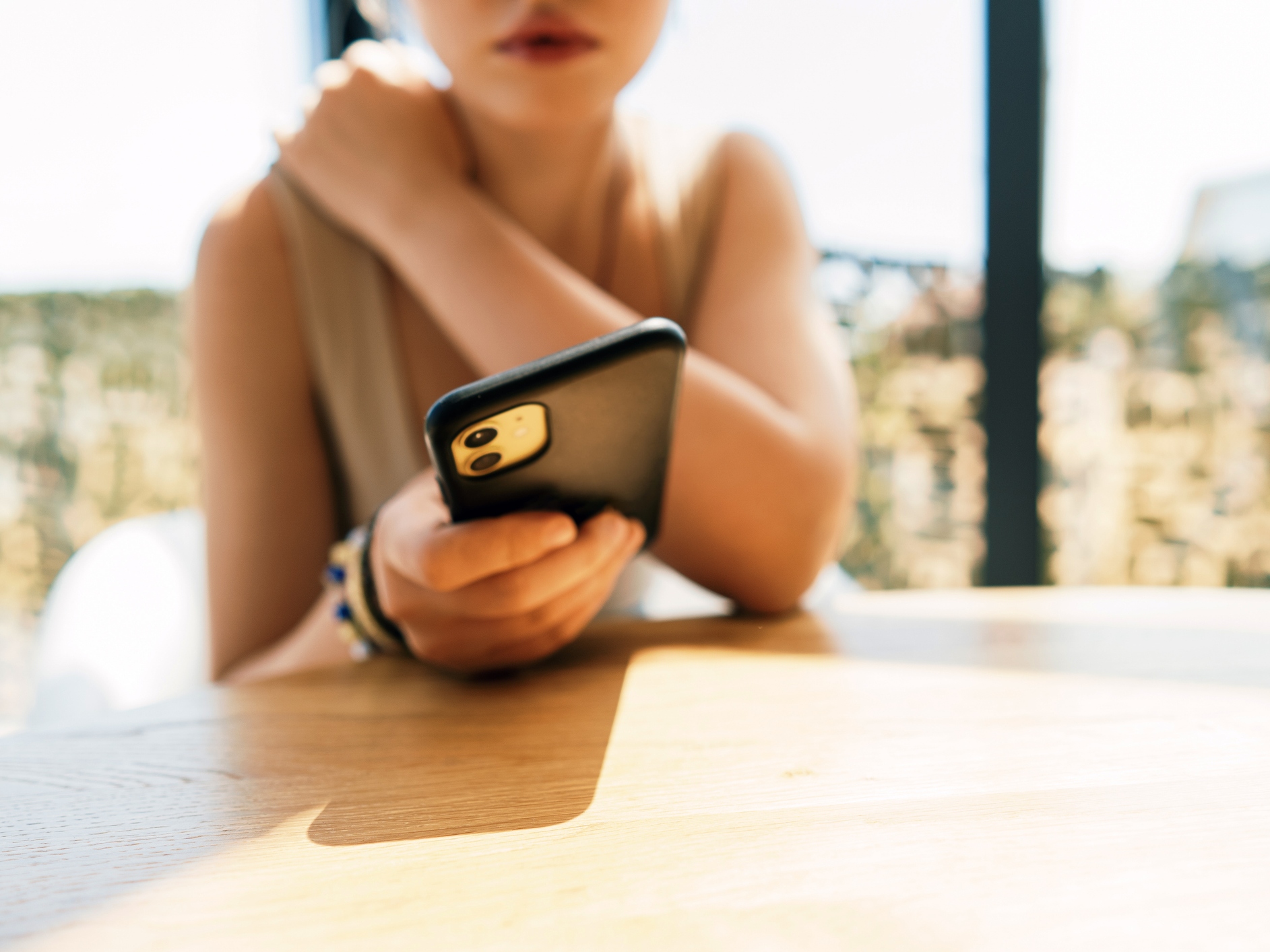 caption: A young woman sits at a sunlit table, holding a smartphone with a soft focus background. The scene conveys casual connectivity, focus on the device, and a relaxed moment of everyday life.