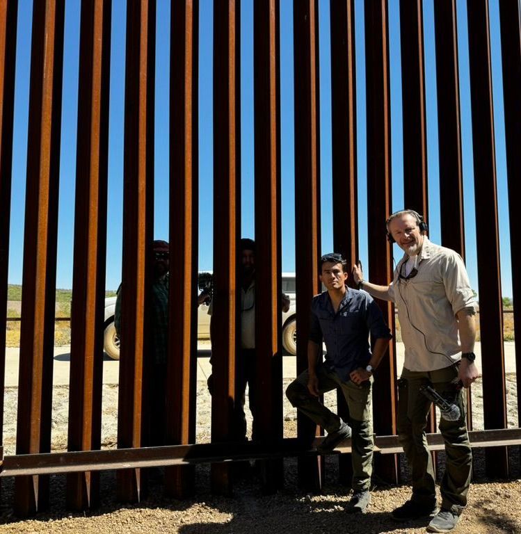 caption: Host of 'The Wild' Chris Morgan (right) stands with biologist Ganesh Marin (left) in front of the 30-foot border wall along the US/Mexico border.