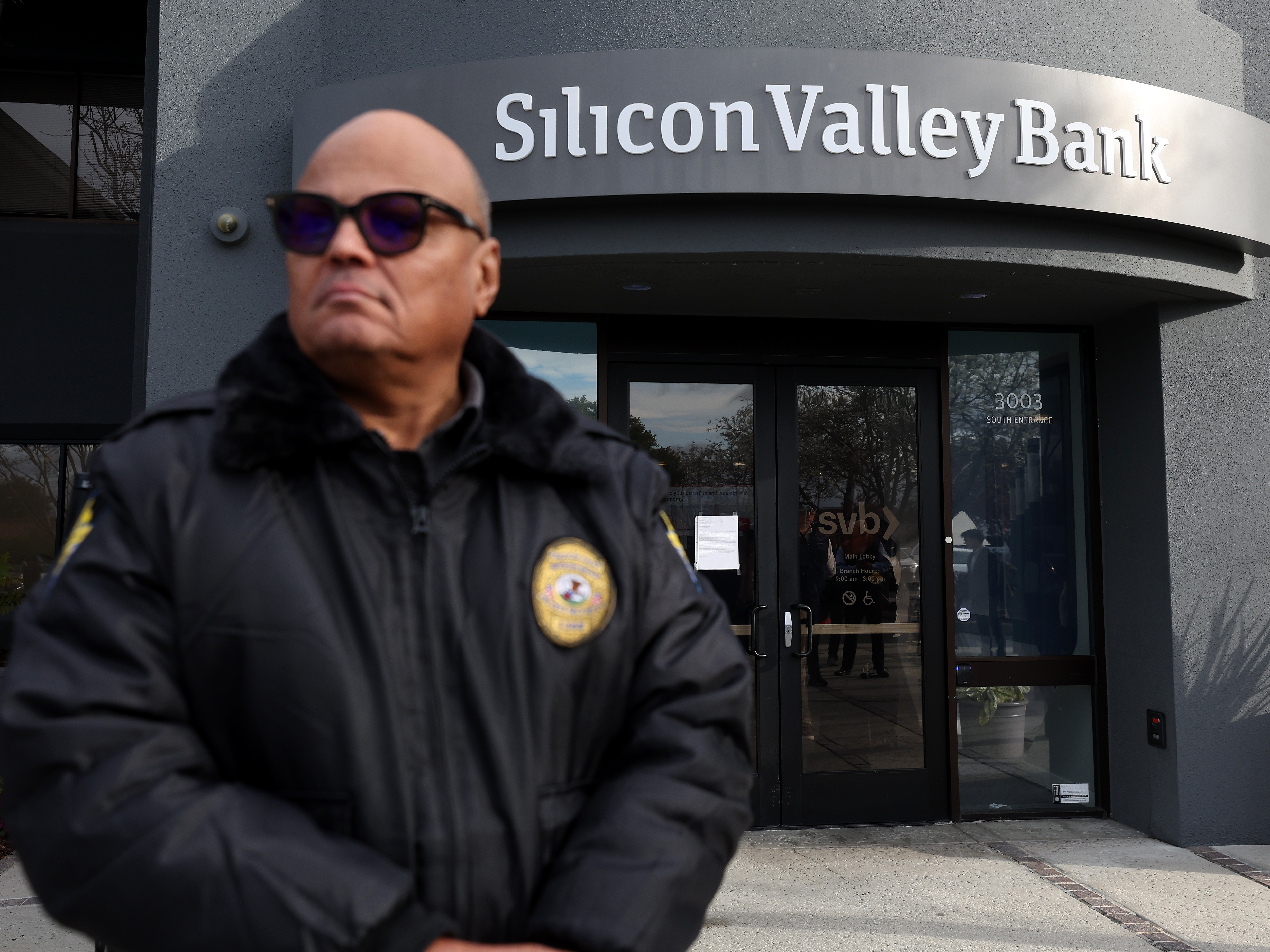 caption: A security guard at Silicon Valley Bank monitors a line of people outside the office in Santa Clara, Calif., on March 13, 2023. The Fed will now need to factor in the impact of the collapse of the lender, along with Signature Bank, as it makes its next decision on interest rates.