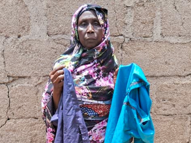 caption: Mariam Mohammed, a widow, stands outside her home in Bama, Nigeria. She's holding her younger son Babagana's favorite clothes. He died in early February from complications of sickle cell disease. She had taken him to a U.S.-funded clinic for treatment but at that time the facility was shuttered due to a stop-work order issued by the Trump Administration.