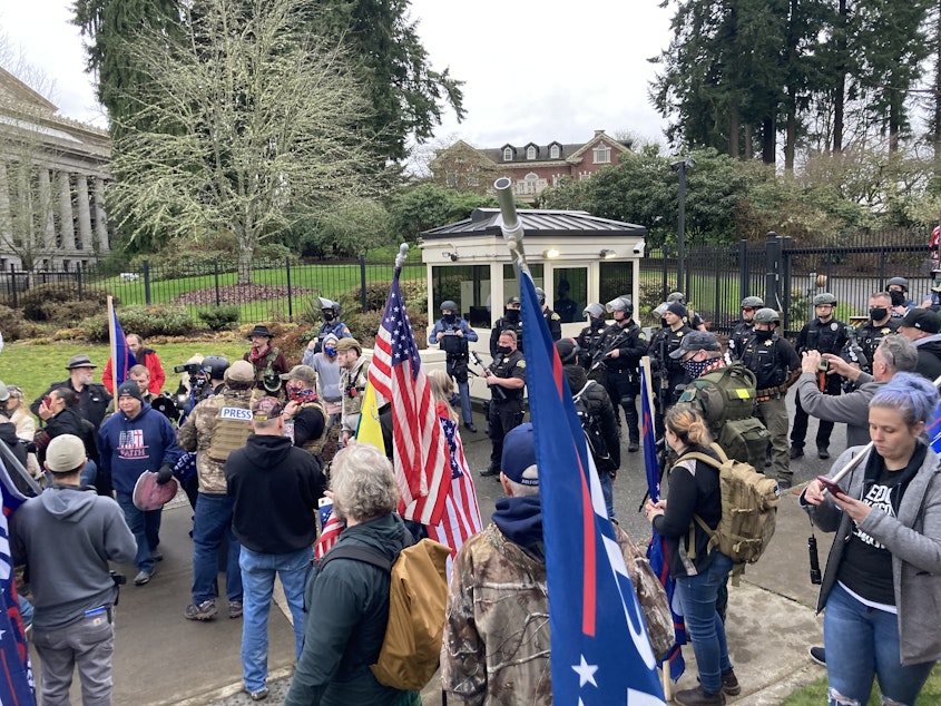 caption: Pro-Trump supporters and riot-clad officers gather at the gates to the governor's residence in Olympia on January 6, 2021. This photograph was taken following a security breach during which a large crowd pushed through a gate and swarmed the lawn of the residence. Gov. Jay Inslee on Thursday cited that incident and the attack on the U.S. Capitol that same day as evidence that democracy is under attack in the United States.