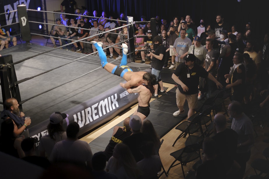 caption: Titus Alexander flies off the top rope into his opponent as the crowd is moved away by security.