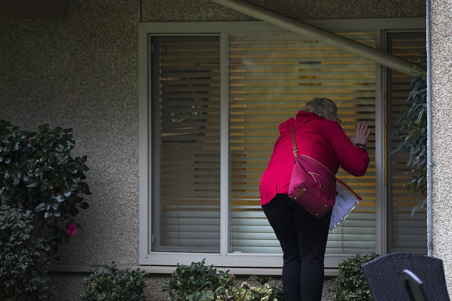 caption: A woman waves after knocking on her mother's window at the Life Care Center of Kirkland, the long-term care facility at the epicenter of the coronavirus outbreak in Washington state, on Monday, March 2, 2020, in Kirkland.