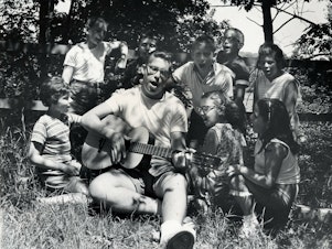 caption: A camp counselor plays the guitar and leads children in singing at Camp Butwin in Minnesota in 1962.