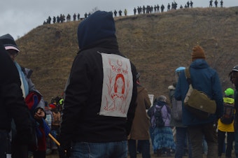 caption: A demonstrator faces police officers at the top of a hill containing Lakota burial sites. His sweatshirt says "water is life" in DinÃ©, the Navajo language.