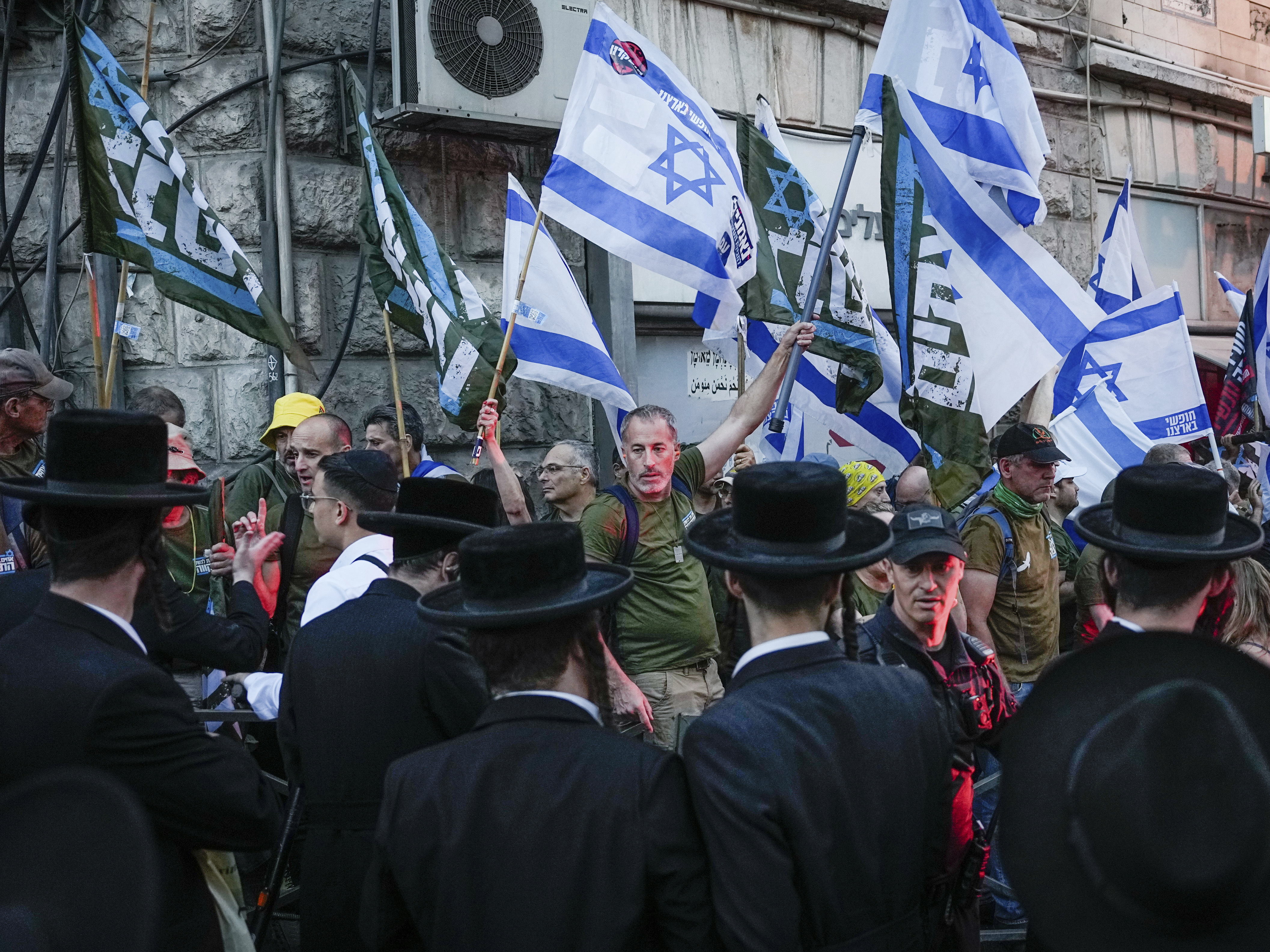 caption: Members of the 'Brothers in Arms' reservist protest group wave Israeli flags during a demonstration in the ultra-Orthodox neighborhood of Mea Shearim, demanding equality in Israel's military service, in Jerusalem on Sunday, March 31, 2024.