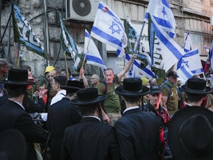 caption: Members of the 'Brothers in Arms' reservist protest group wave Israeli flags during a demonstration in the ultra-Orthodox neighborhood of Mea Shearim, demanding equality in Israel's military service, in Jerusalem on Sunday, March 31, 2024.