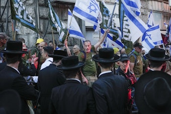caption: Members of the 'Brothers in Arms' reservist protest group wave Israeli flags during a demonstration in the ultra-Orthodox neighborhood of Mea Shearim, demanding equality in Israel's military service, in Jerusalem on Sunday, March 31, 2024.
