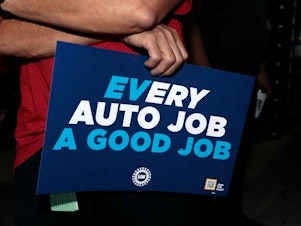 caption: UAW supporters and members hold signs outside the Stellantis Assembly Plant in Sterling Heights, Mich., on July 12, 2023. Electric vehicles may not be at the heart of the contract talks between the UAW and the Detroit automakers, but as the sign subtly shows, they loom large in the negotiations.