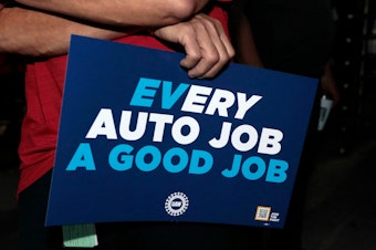 caption: UAW supporters and members hold signs outside the Stellantis Assembly Plant in Sterling Heights, Mich., on July 12, 2023. Electric vehicles may not be at the heart of the contract talks between the UAW and the Detroit automakers, but as the sign subtly shows, they loom large in the negotiations.