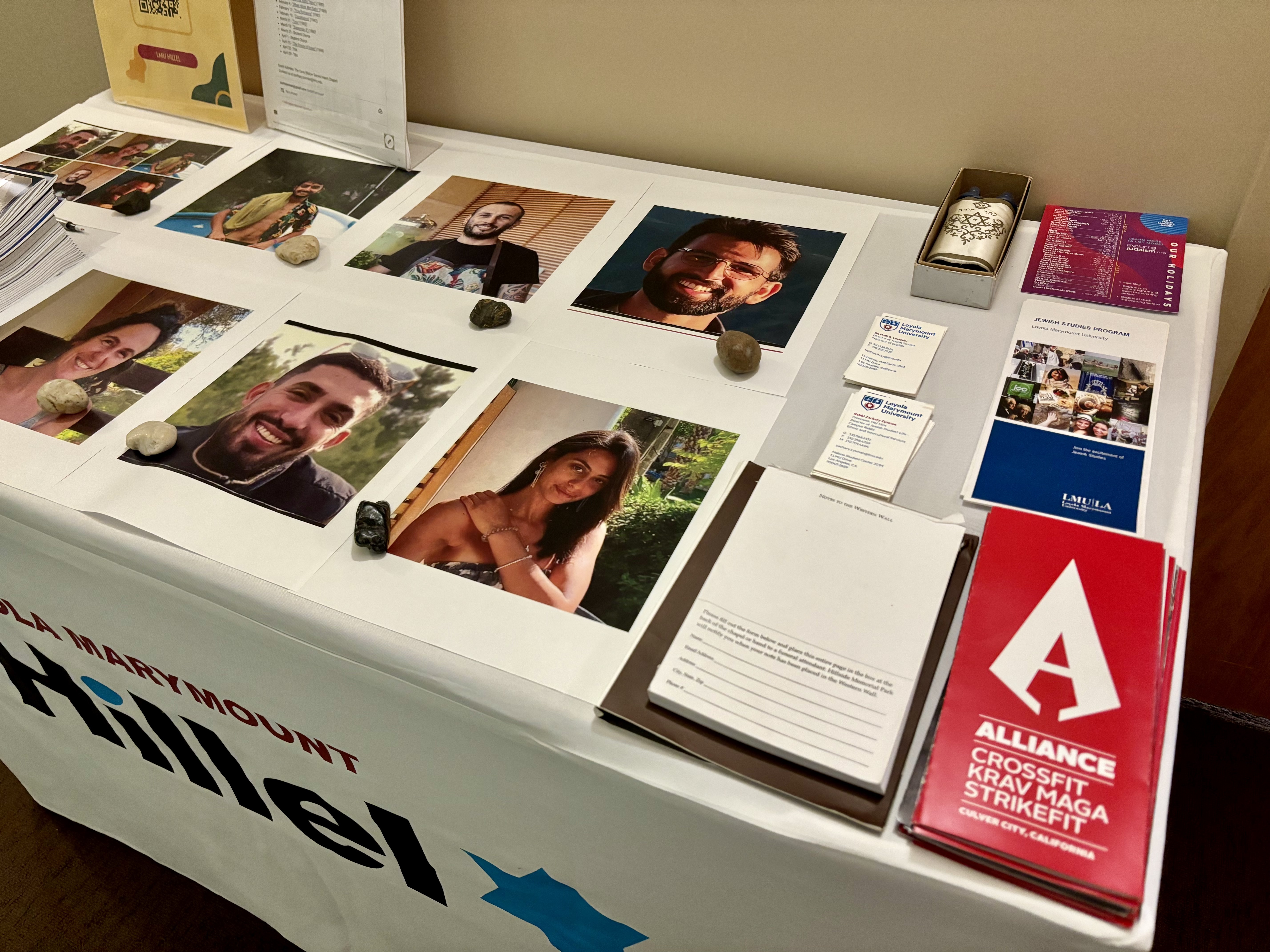 caption: A table covered with a cloth featuring the Loyola Marymount Hillel logo in the front, and six portraits of hostages who were killed sitting on top of it.