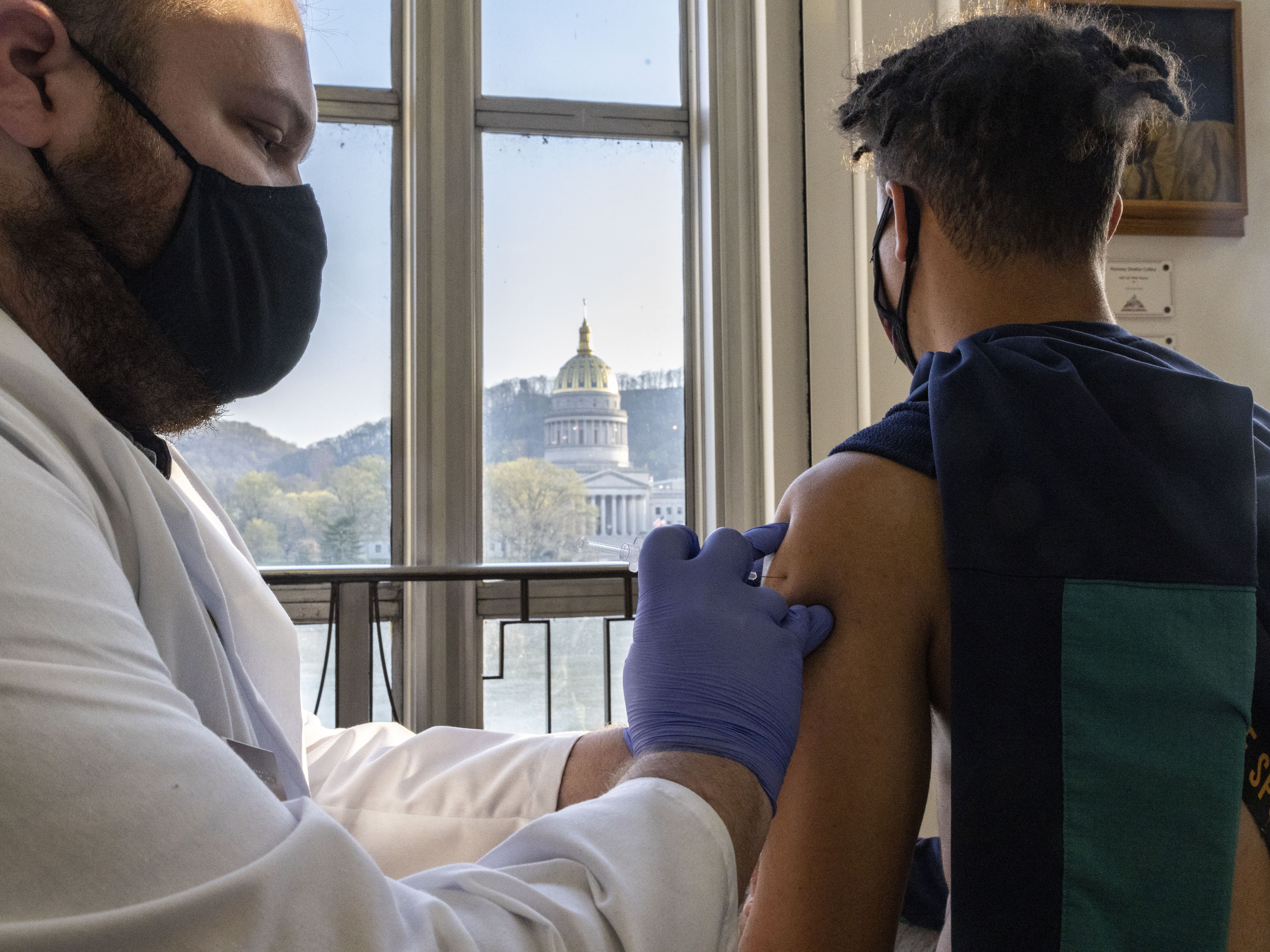 caption: A young man receives a COVID-19 vaccine in Charleston earlier this month while overlooking the West Virginia Capitol Building. The state recently announced a plan to give $100 savings bonds to people between the ages of 16 and 35 who get vaccinated.