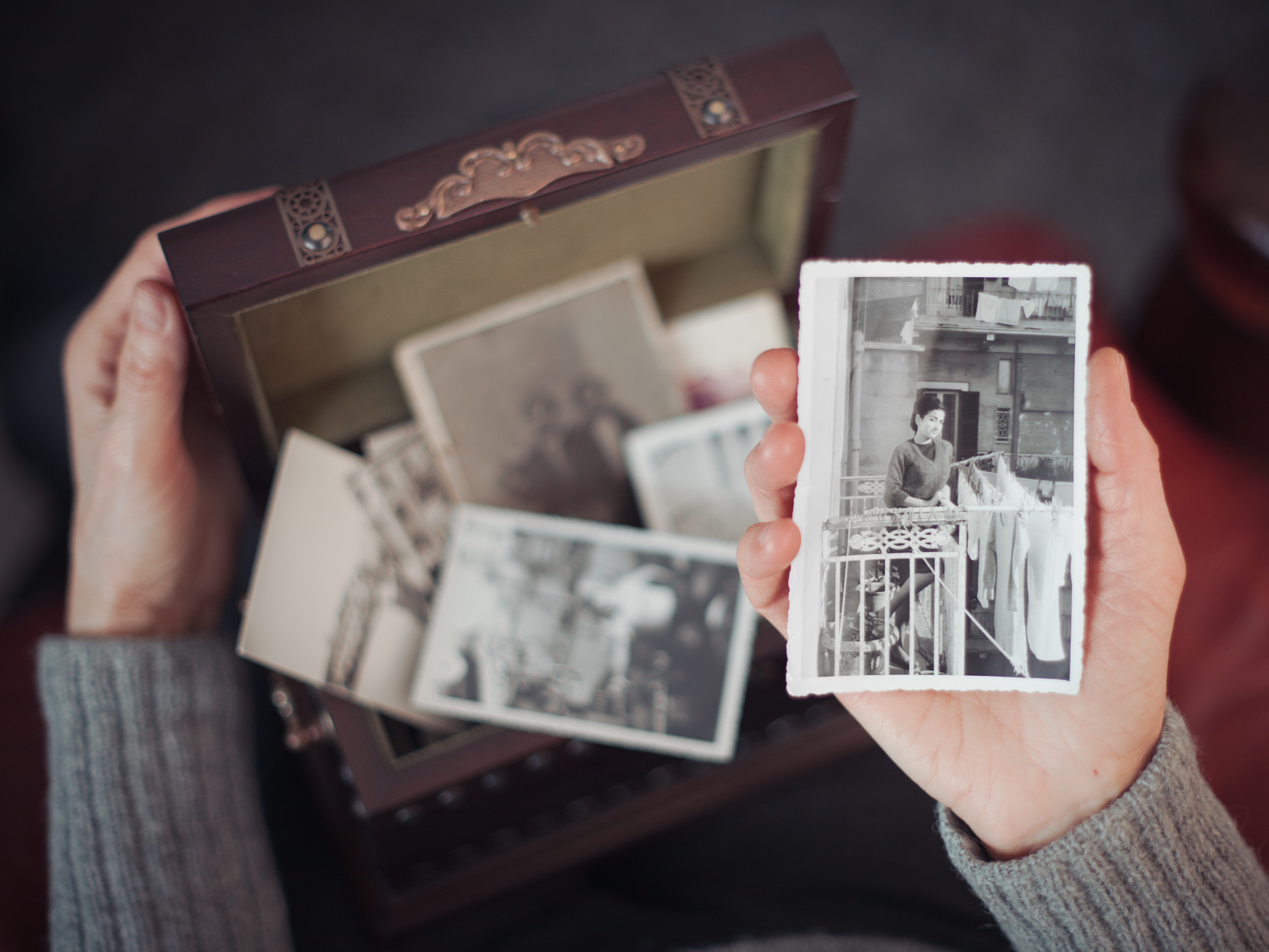 Hands of woman discovering a treasure chest full of photographs and holding an old black and white photograph of a smiling woman standing on a balcony in Milan in 1960s.