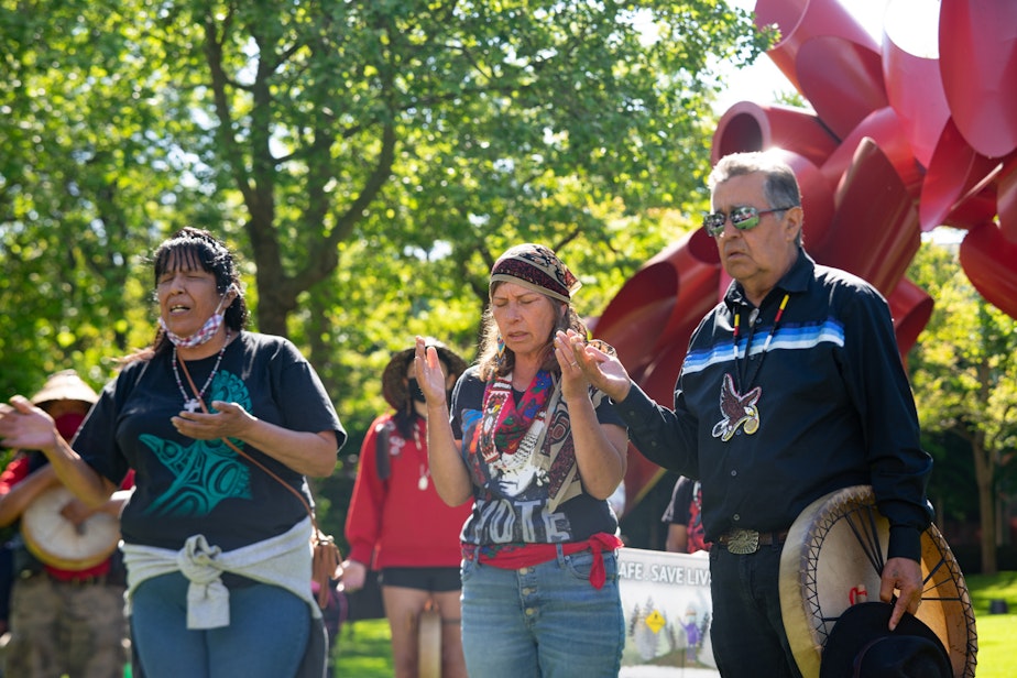 caption: From left, Triva Sampson, Siamel’wit, and Lummi Carver Douglas (Sit-ki-kadem) James sing and pray near the 24-foot totem pole on Saturday, May 22, 2021, at Seattle Center.