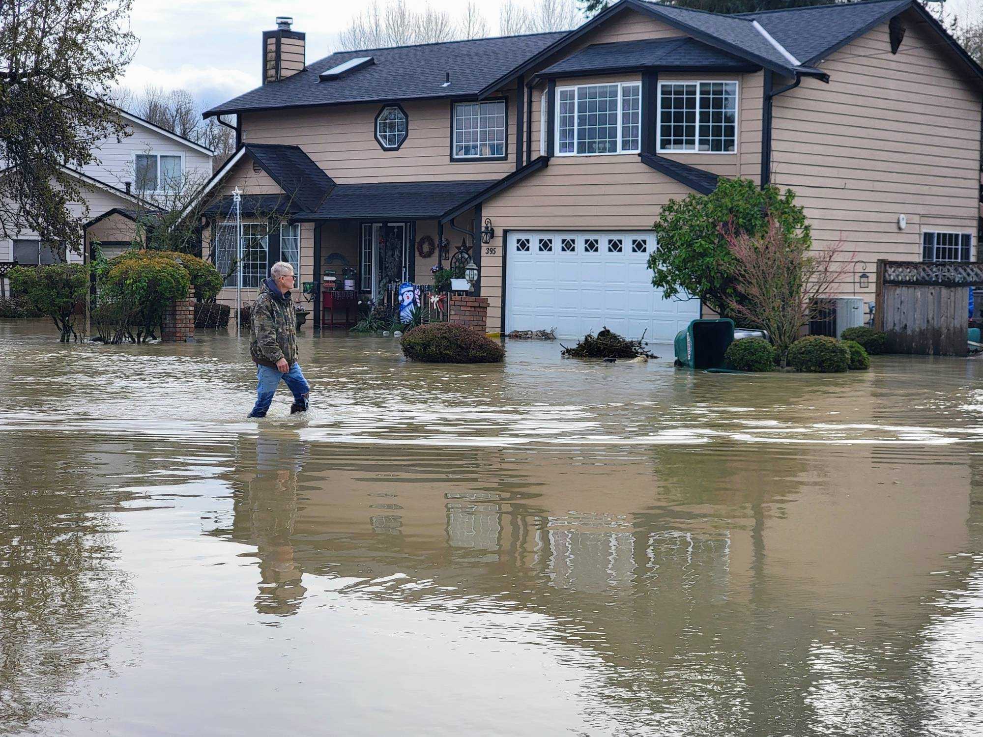 caption: Dean Johnson wades through floodwaters outside his home in Pacific on Dec. 16, 2025, after saving his chickens from a flooded coop.