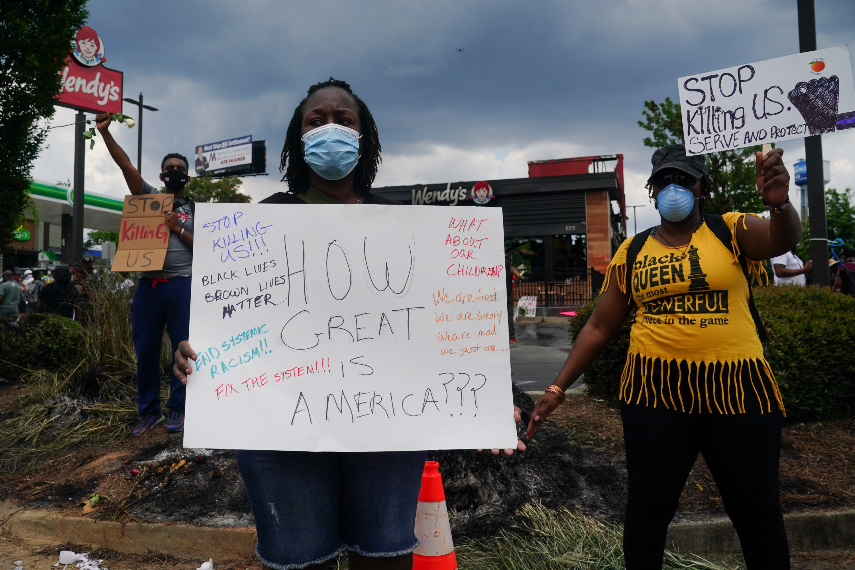 caption: People hold signs toward traffic outside a burned Wendys restaurant on the second day following the police shooting death of Rayshard Brooks in the restaurant parking lot June 14, 2020, in Atlanta, Georgia. (ELIJAH NOUVELAGE/AFP via Getty Images)