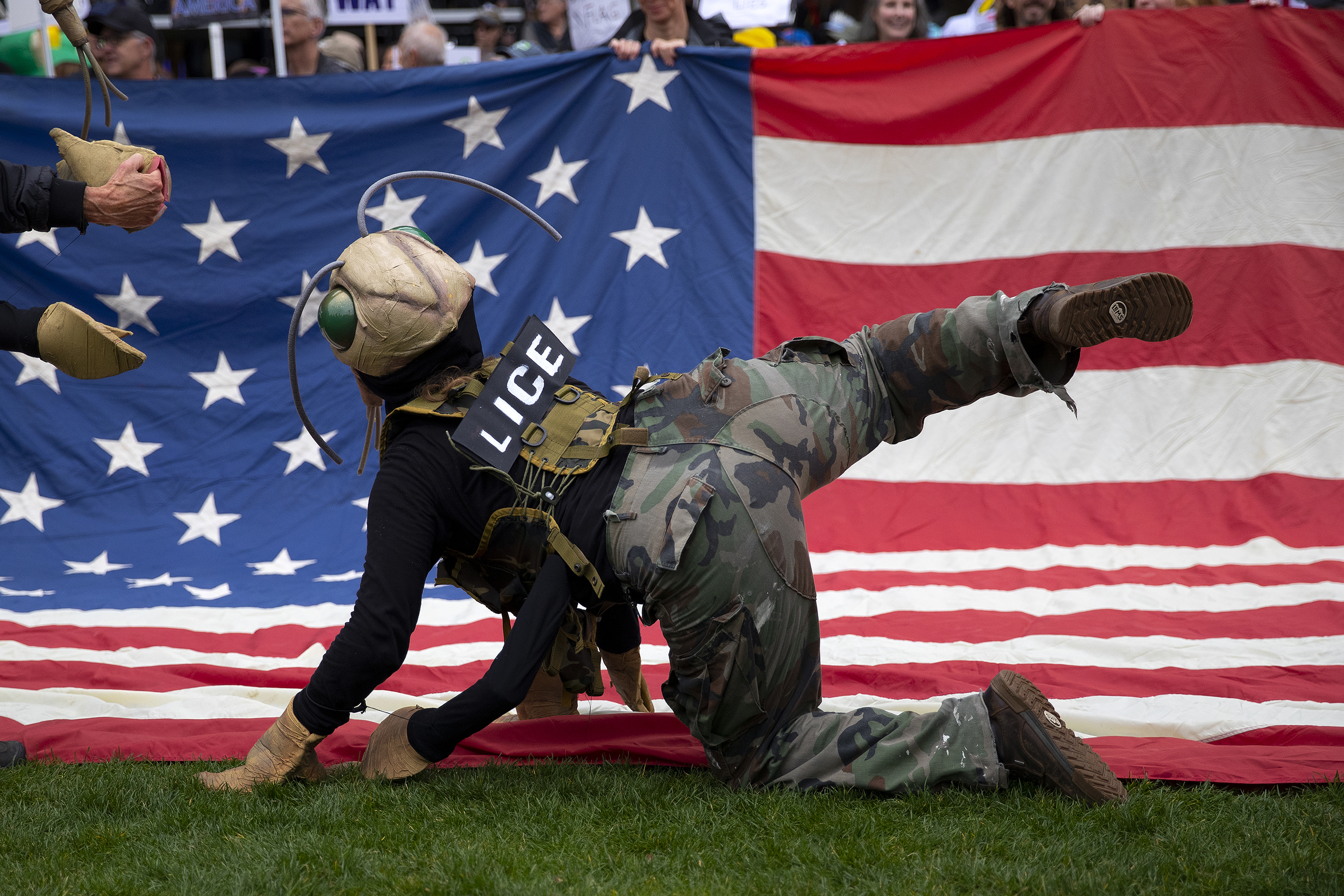 caption: A protester pretends to pee on an American flag dressed as ‘lice’ during the No Kings rally and march on Saturday, October 18, 2025, at Seattle Center. 