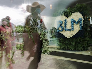 caption: People are reflected in a window near a flower arrangement that includes the acronym for Black Lives Matter as they wait in line to attend the public viewing for George Floyd at the Fountain of Praise church on June 8, 2020 in Houston.