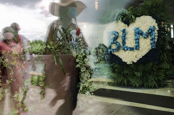 caption: People are reflected in a window near a flower arrangement that includes the acronym for Black Lives Matter as they wait in line to attend the public viewing for George Floyd at the Fountain of Praise church on June 8, 2020 in Houston.