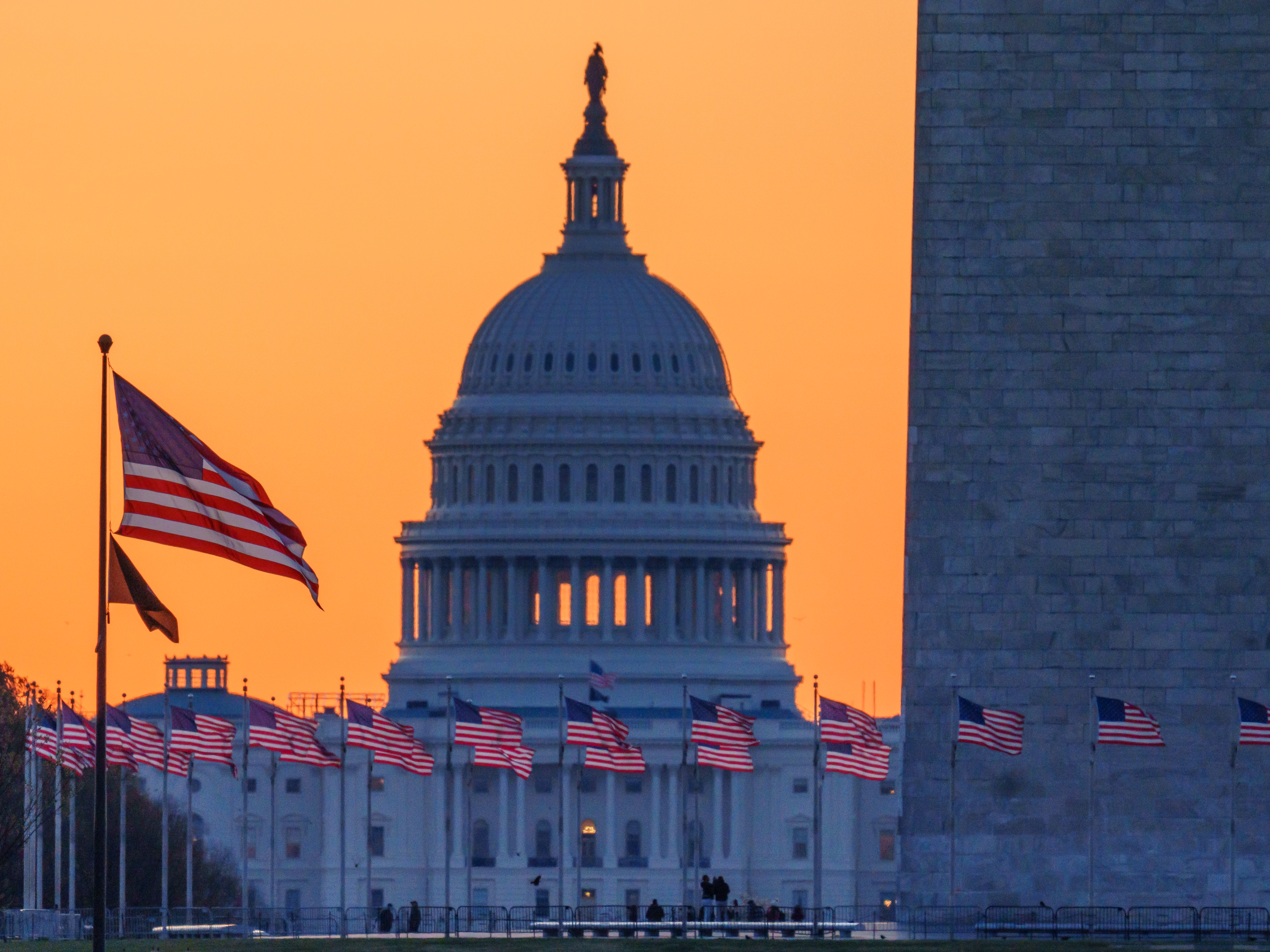 caption: Sunrise turns the sky orange behind the U.S. Capitol building as it illuminates the U.S. flags circling the base of the Washington Monument on March 23 in Washington, D.C.