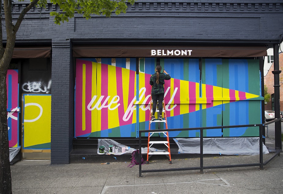 caption: Sara Thompson paints a mural on Sunday, May 3, 2020, at the intersection of East Pike Street and Belmont Avenue in Seattle. When complete, the mural will read "United we stand, divided we fall."