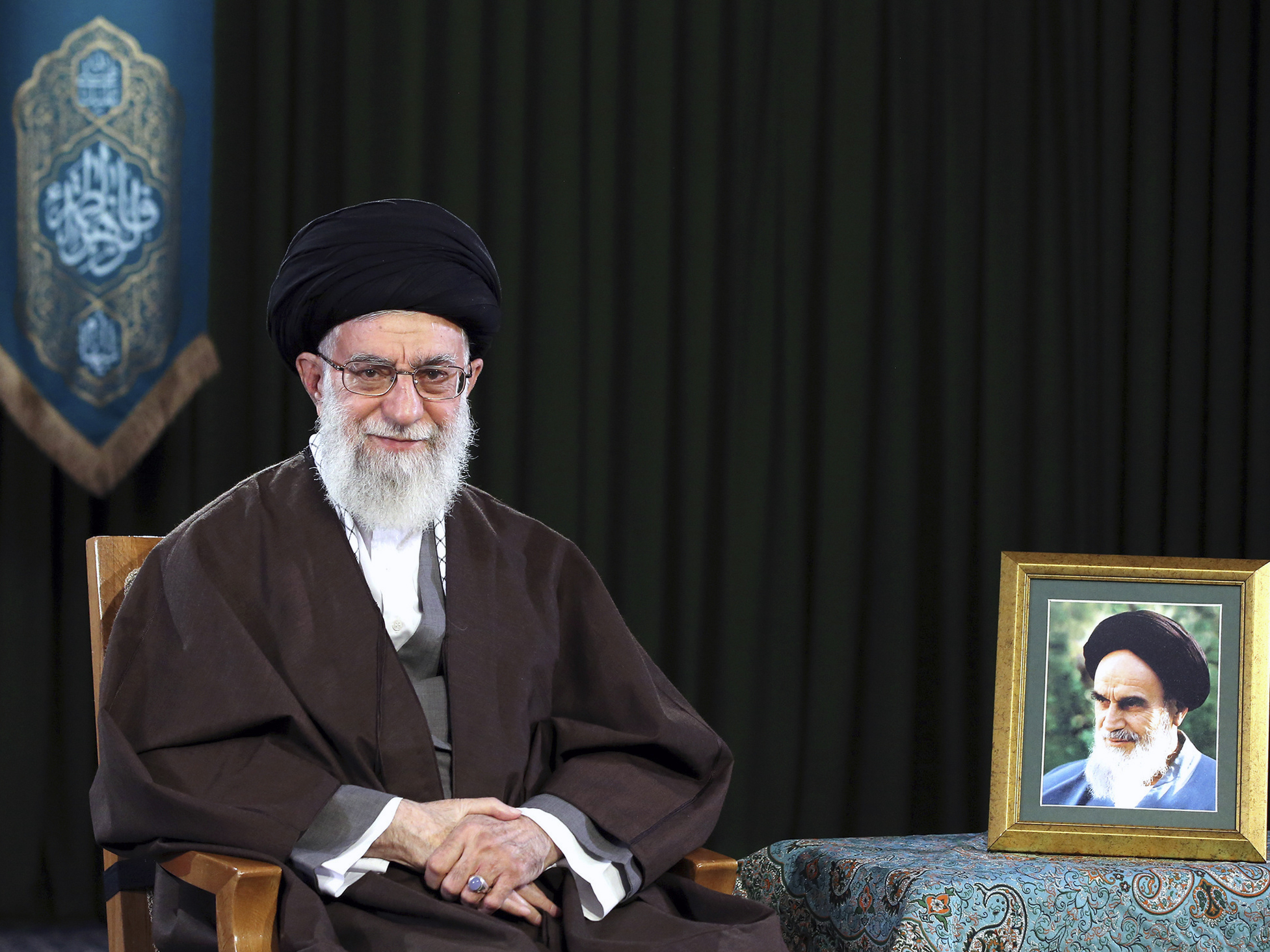 caption: In this 2017 photo, Ayatollah Ali Khamenei, Iran's supreme leader, sits in a session to deliver his message for the Iranian New Year. A portrait of the late revolutionary founder, Ayatollah Ruhollah Khomeini, is next to him.
