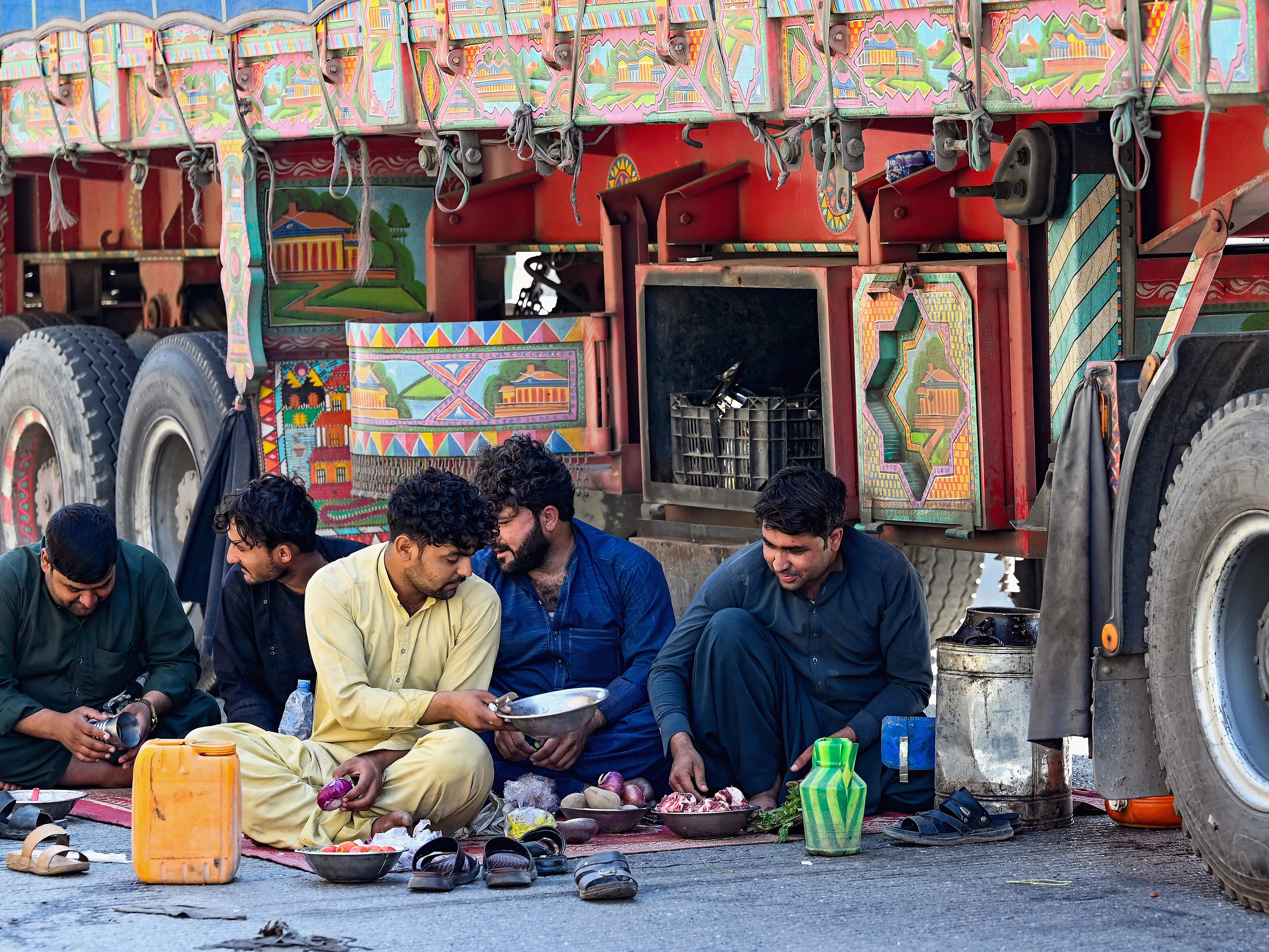 caption: Truck drivers eat on the ground by their stalled vehicles near Torkham, Pakistan, on Oct. 13, 2025. More than three months later, the Torkham border remains closed with no end in sight.