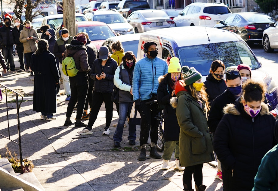 caption: City residents wait in a line extending around the block to receive free at-home rapid COVID-19 test kits in Philadelphia on Monday.